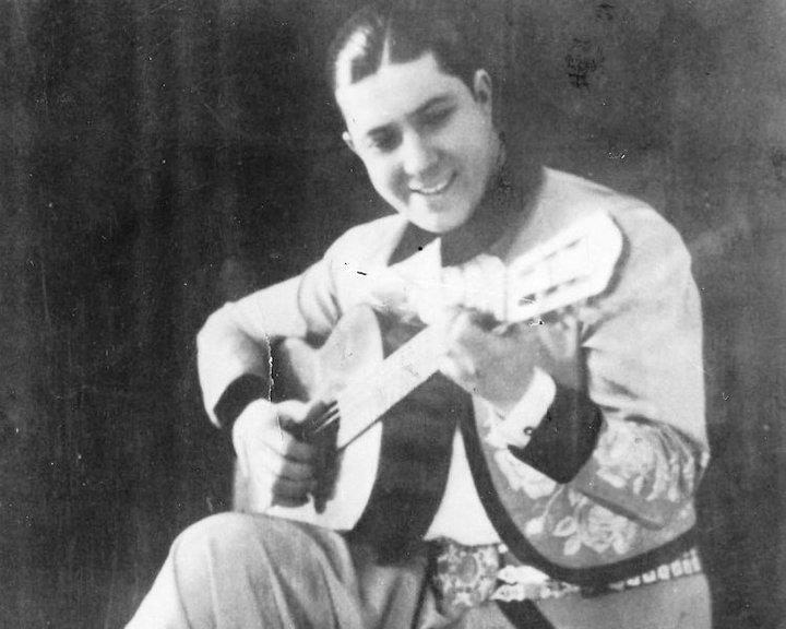 Carlos Gardel sits holding an acoustic guitar in a black and white archival photograph.
