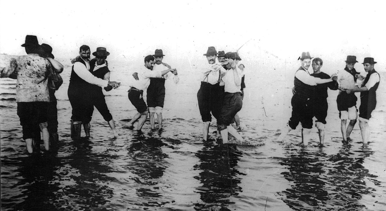 Historical black and white photograph of men dancing in shallow water by the Río de la Plata.